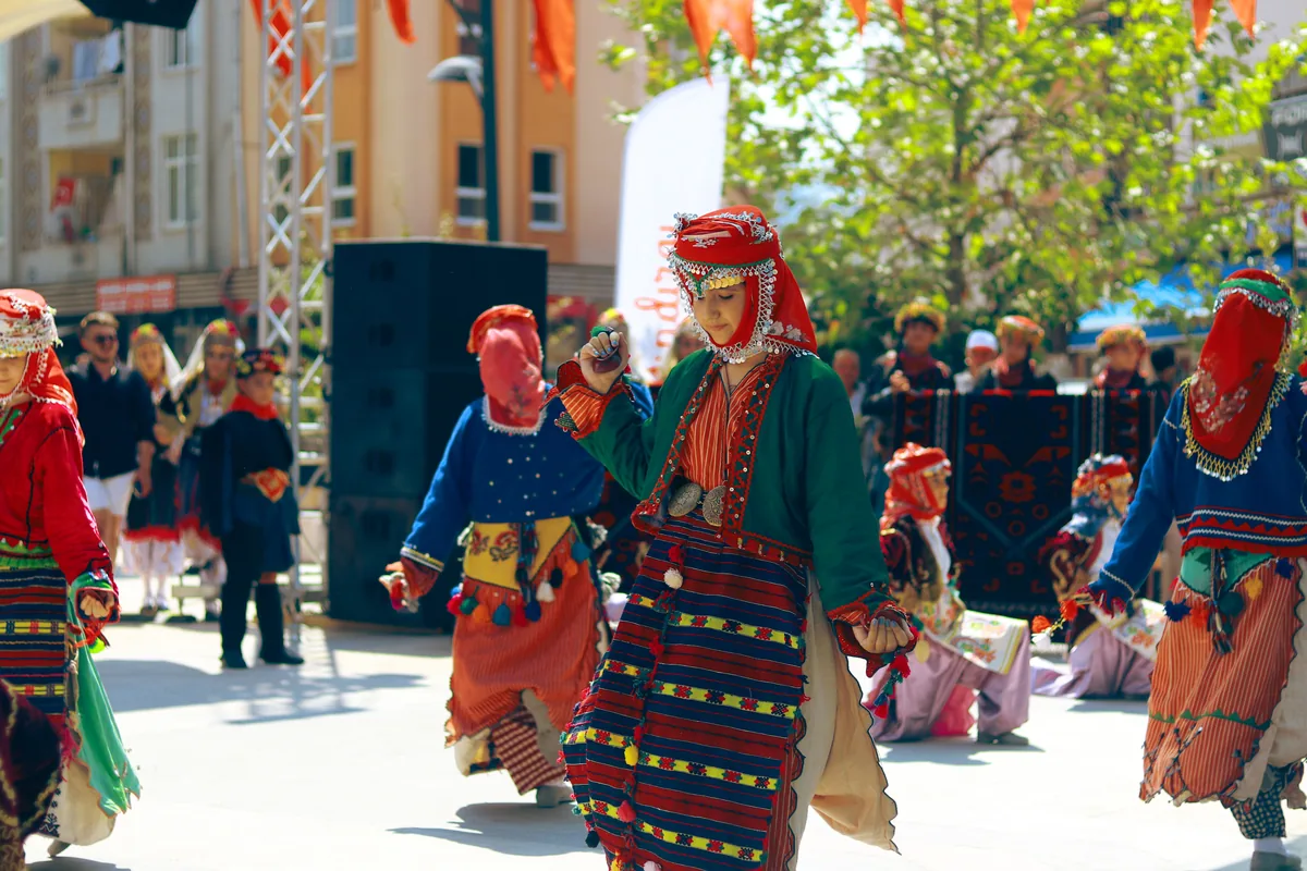Tanečníci v pruhovaných, vyšívaných regionálních krojích s červenými šátky se pohybují v řadě po ulici na venkovním festivalu, v pozadí je pódium a přihlížející