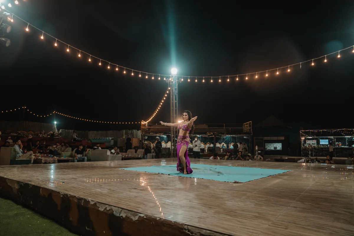 A belly dancer in a magenta costume on a small turquoise rug at the centre of a wide wooden stage, under arcs of string lights, with seated diners around the edge