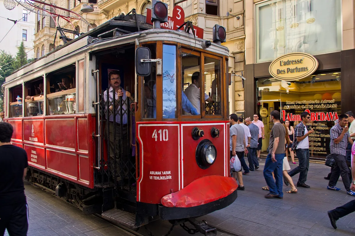 Um elétrico histórico vermelho, o número 410, com o condutor à porta aberta, numa movimentada rua pedonal ladeada de antigas fachadas de pedra