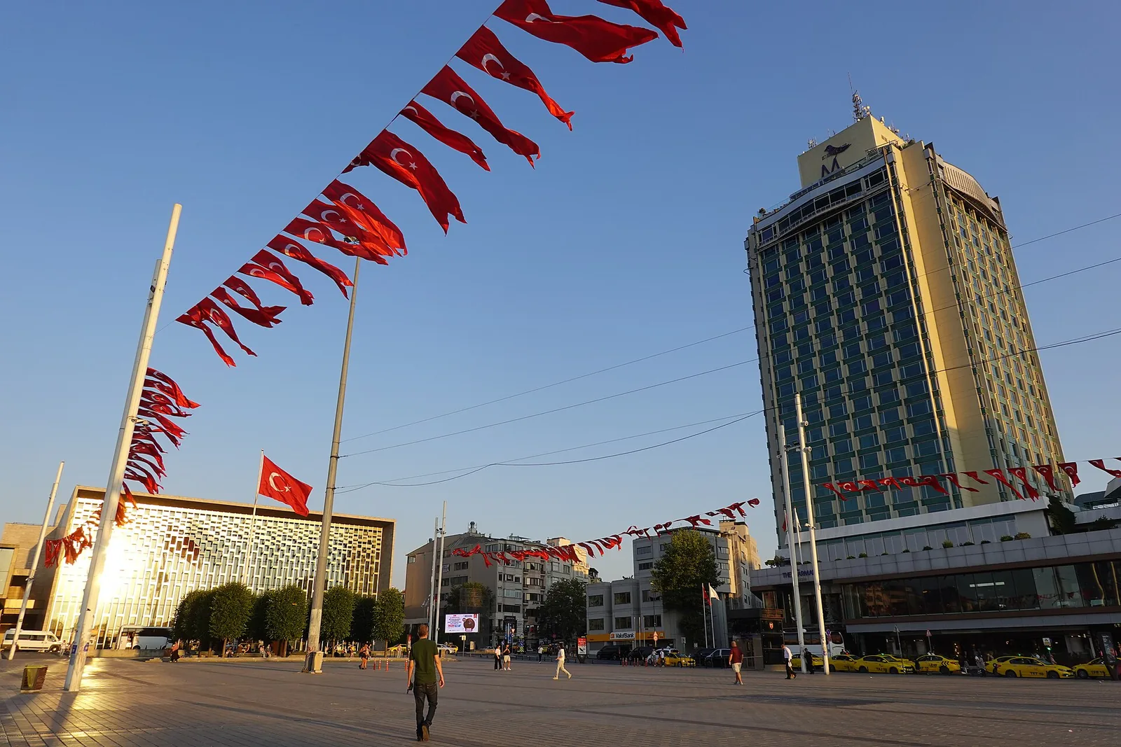 Taksim-pladsen på en solrig dag, med et moderne hotelhøjhus, der rejser sig bag flagranker, og mennesker, der krydser den brolagte plads